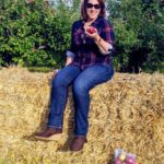 visual image of a woman wearing a plaid shirt and jeans, sitting on a hay bale in an orchard with an apple in her hand and a bag of apples on the ground beside her 
